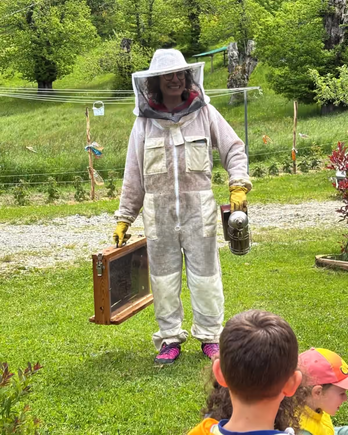 Apicoltrice in tuta con un telaio in mano davanti a un gruppo di bambini, in un prato